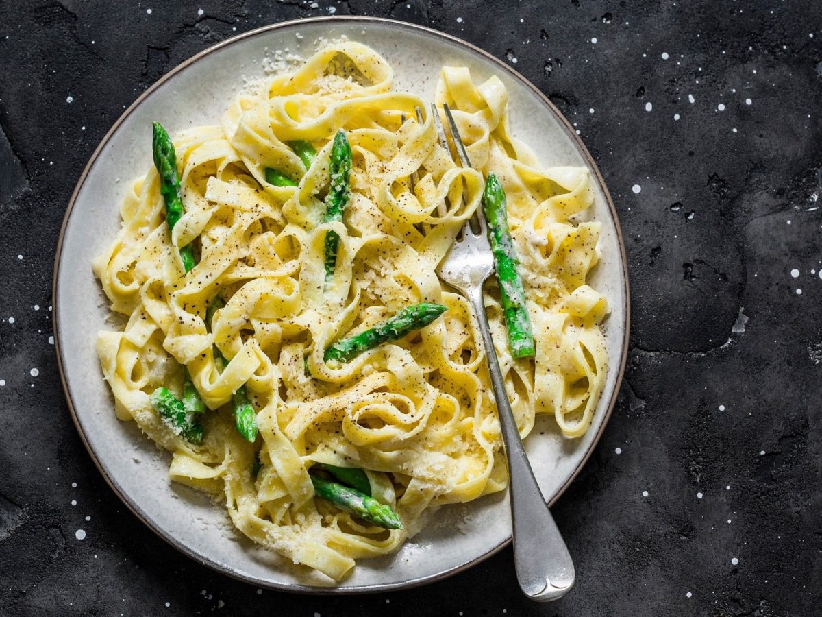 Frischkäse-Pasta mit grünem Spargel auf einem Teller, darauf eine Gabel, auf dunklem Untergrund, Draufsicht.