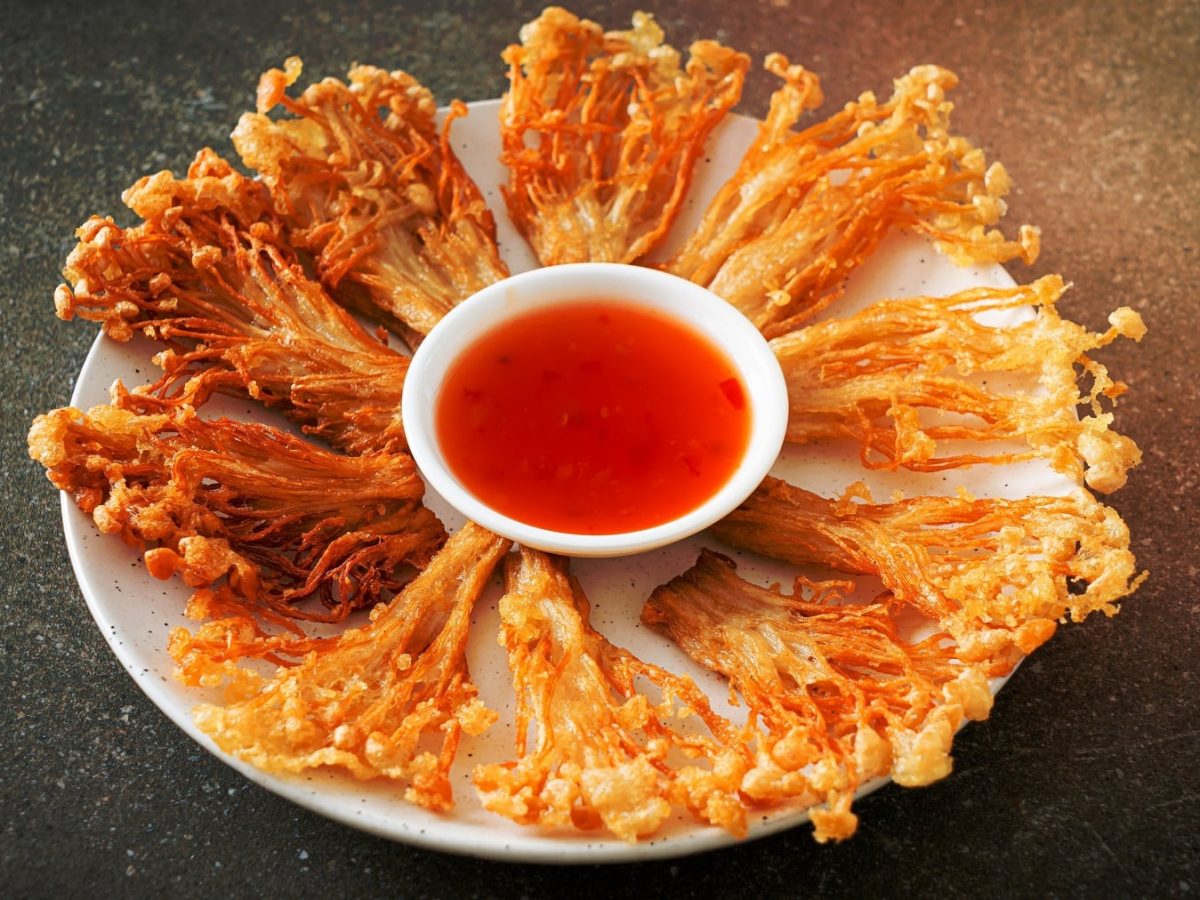 Ein weißer Teller mit den frittierten Enoki und einer kleinen Schale mit Sweet-Chili-Sauce auf einem dunklen Tisch.