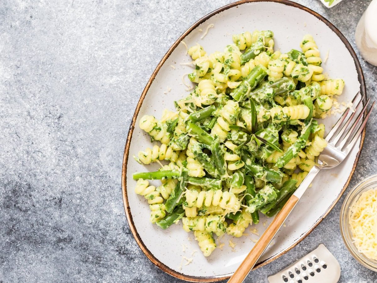 Ein ovaler, weißer Teller mit den Fusilli, grünen Bohnen und Käsesauce. Daneben Rucola, Käse, Salz und Pfeffer.