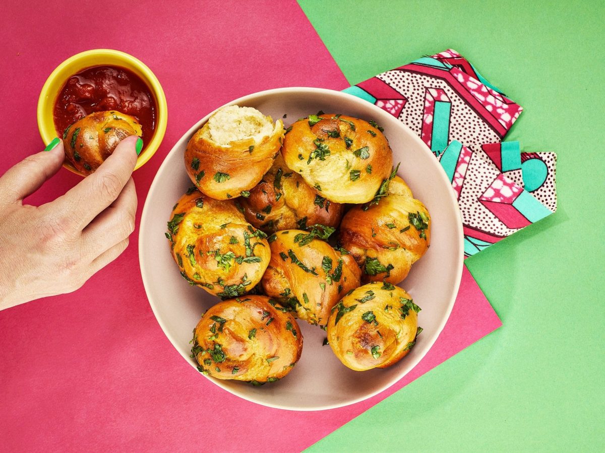 Garlic Knots in einer Schüssel in der Draufsicht, daneben eine Schale mit rotem Dip, in die eine Hand gerade einen der Knoblauchknoten dippt.