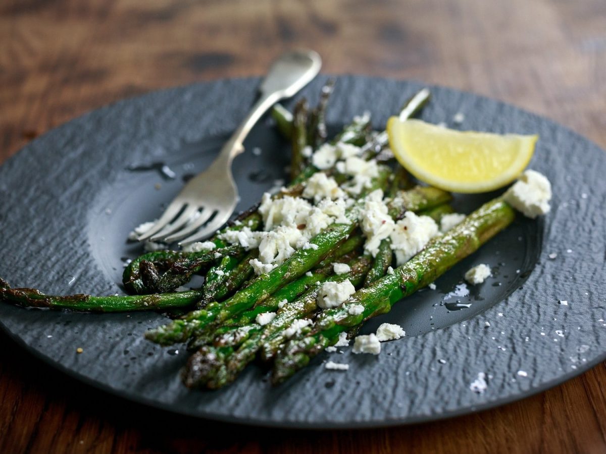 Ein Schiefer-Teller gegrillter Spargel mit Zitrone und Feta, darauf eine Gabel.