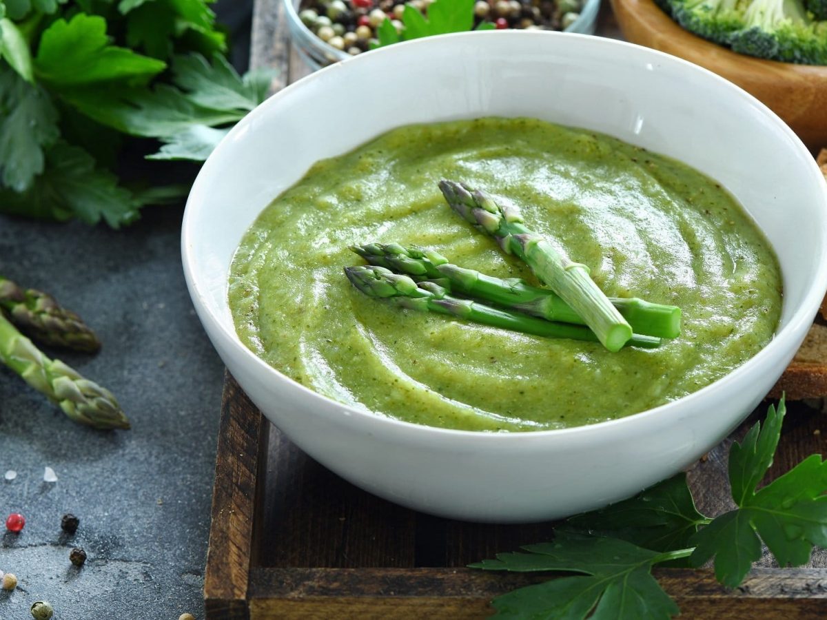 Eine Schüssel grüne Spargelcremesuppe mit Spargelstangen garniert, drumherum liegen Zutaten.