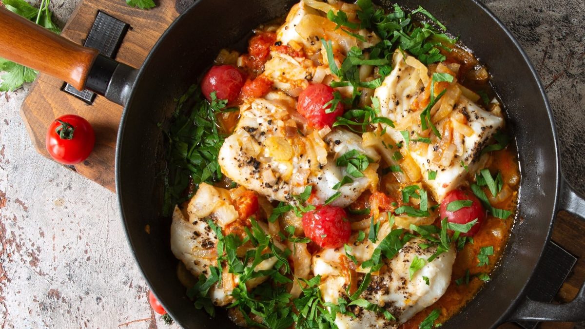 Eine Pfanne mit dem Fisch, Tomaten und Fenchel auf einem Steintisch von oben fotografiert.