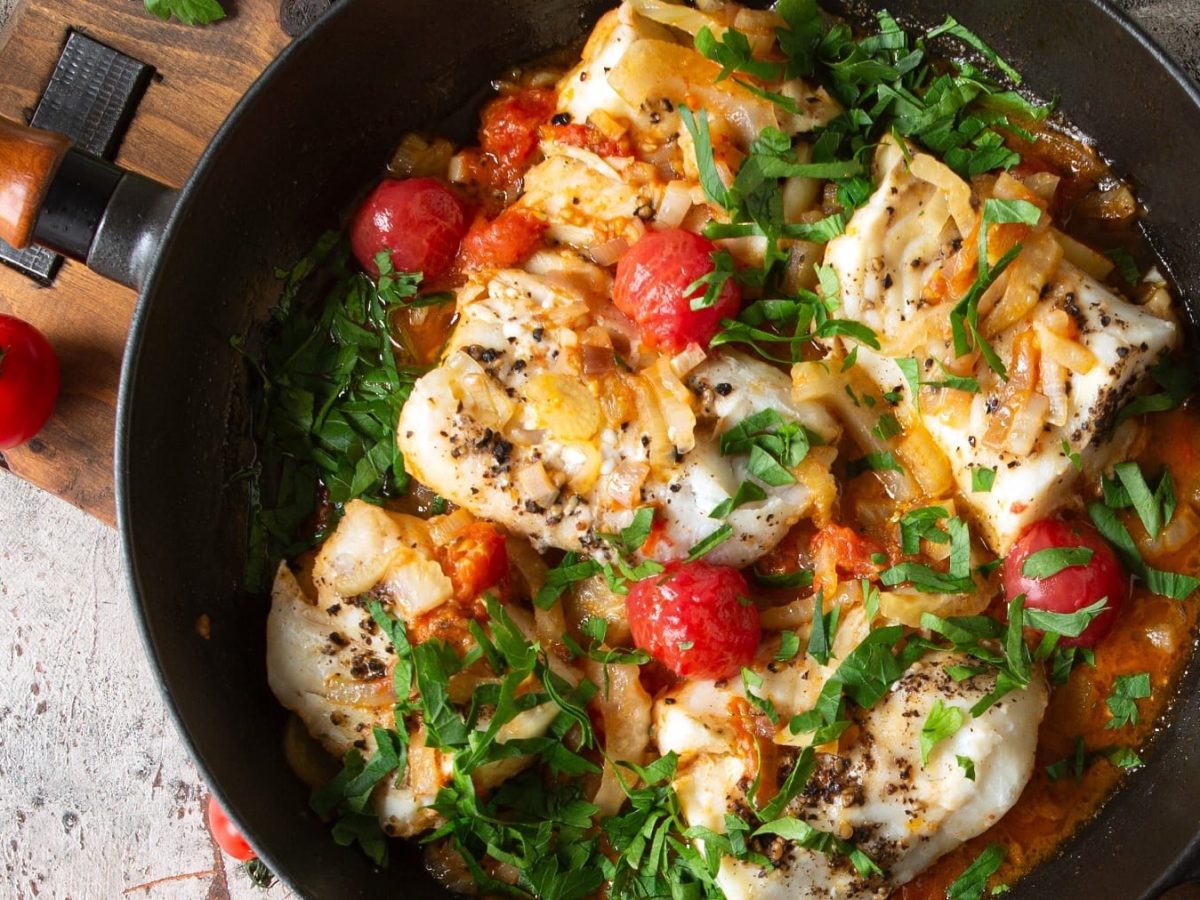 Eine Pfanne mit dem Fisch, Tomaten und Fenchel auf einem Steintisch von oben fotografiert.