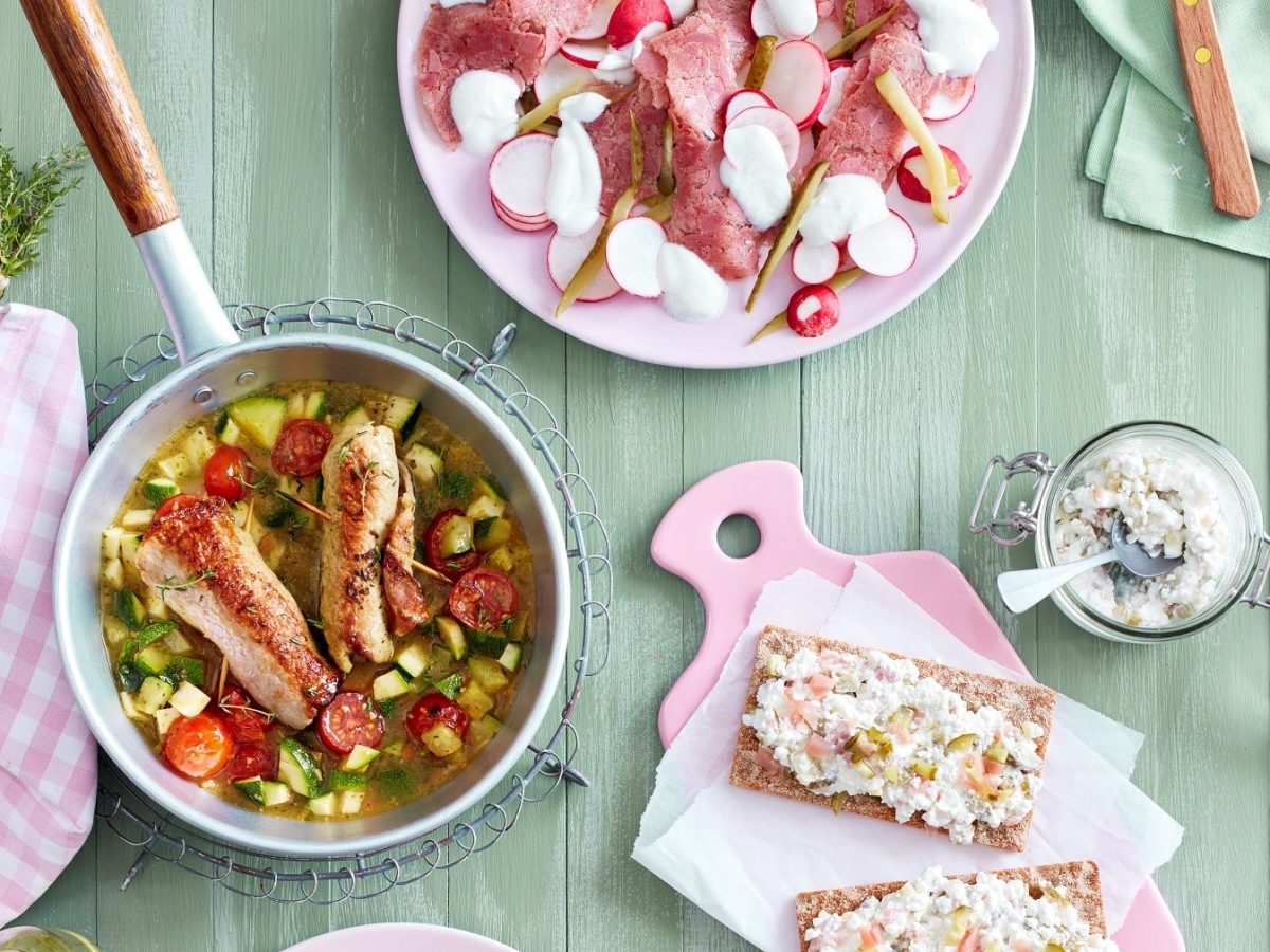 Kalbsröllchen mit Zucchini in einer Pfanne, daneben andere Gerichte, alles in der Draufsicht vor grünem Holzhintergrund.