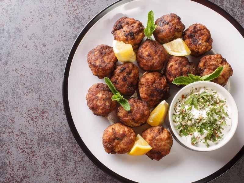 griechische Hackbällchen mit Dip auf weißem Teller, dazu Dip, Kräuter und Zitronenspalten.