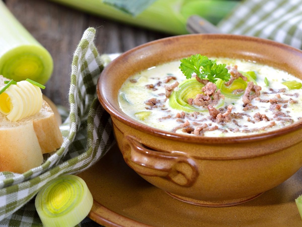 Frontalsicht: In einer runden Tonschale ist eine Hackfleisch-Suppe mit Porree. Drumherum liegen Lauch, Weißbrotscheiben und ein kariertes Küchentuch