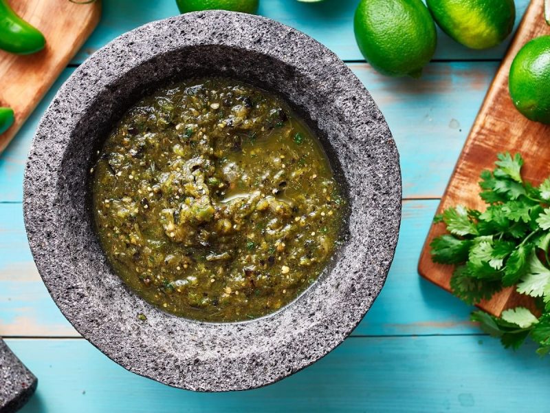 Ein Steintopf mit Limetten-Koriander-Salsa auf einem blauen Tisch neben frischen Zutaten.