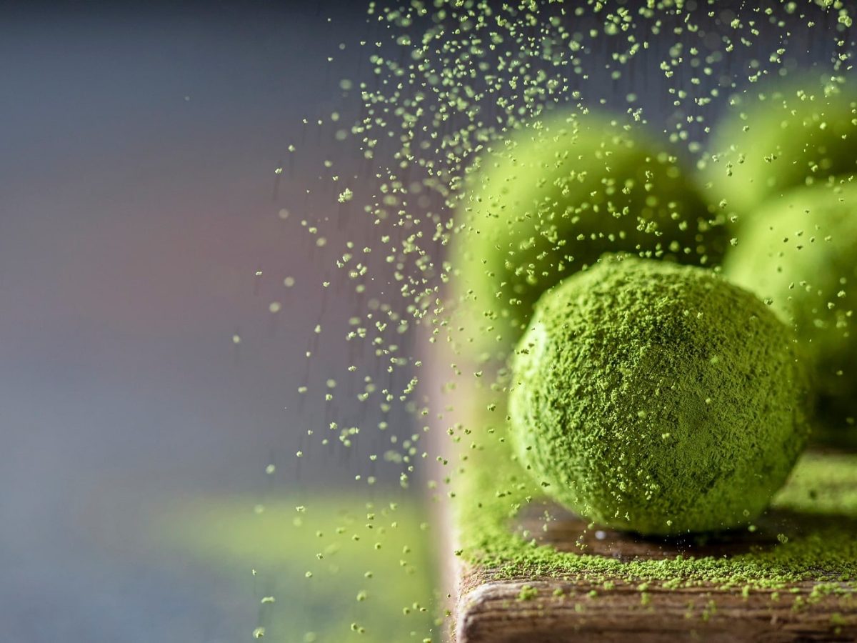 Mehrere Matcha Fudge Bällchen auf einem Holzbrett mit grauem Untergrund. Zudem wird gerade Matcha-Pulver darüber gerieselt.