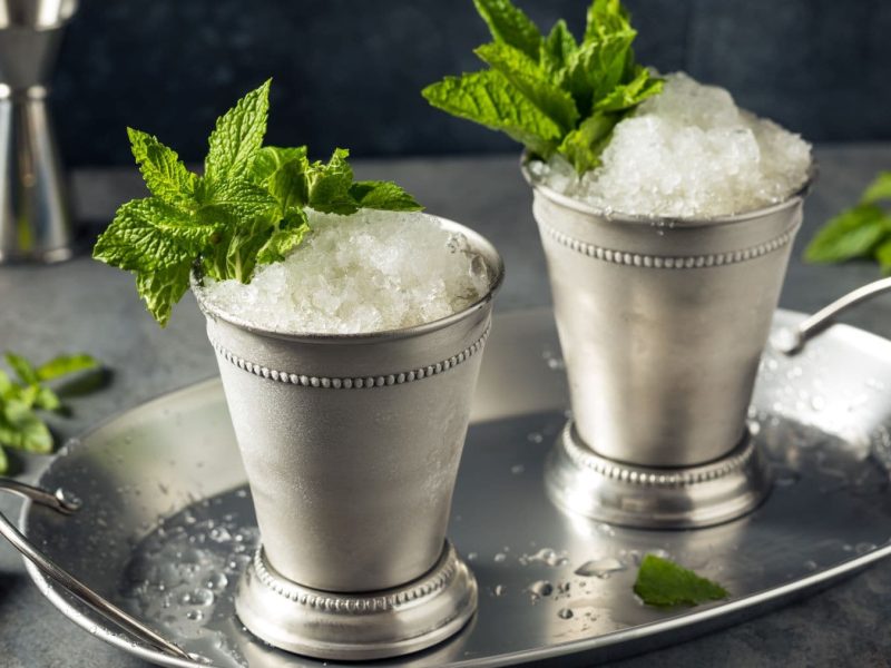 Zwei Mint Julep im Metallbecher auf Metallteller mit Minze. Im Hintergrund Barmaß, Stößel und Minze. Frontalaufnahme.