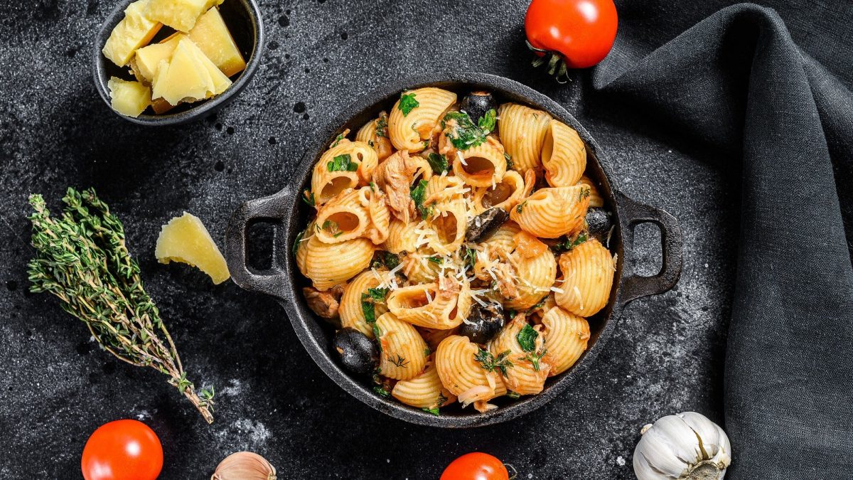 Draufsicht: dunkler Untergund, eine ofenfeste form mit Pasta in Tomaten-Kapern-Sauce. Drum herum liegen Gemüse und Kräuter.