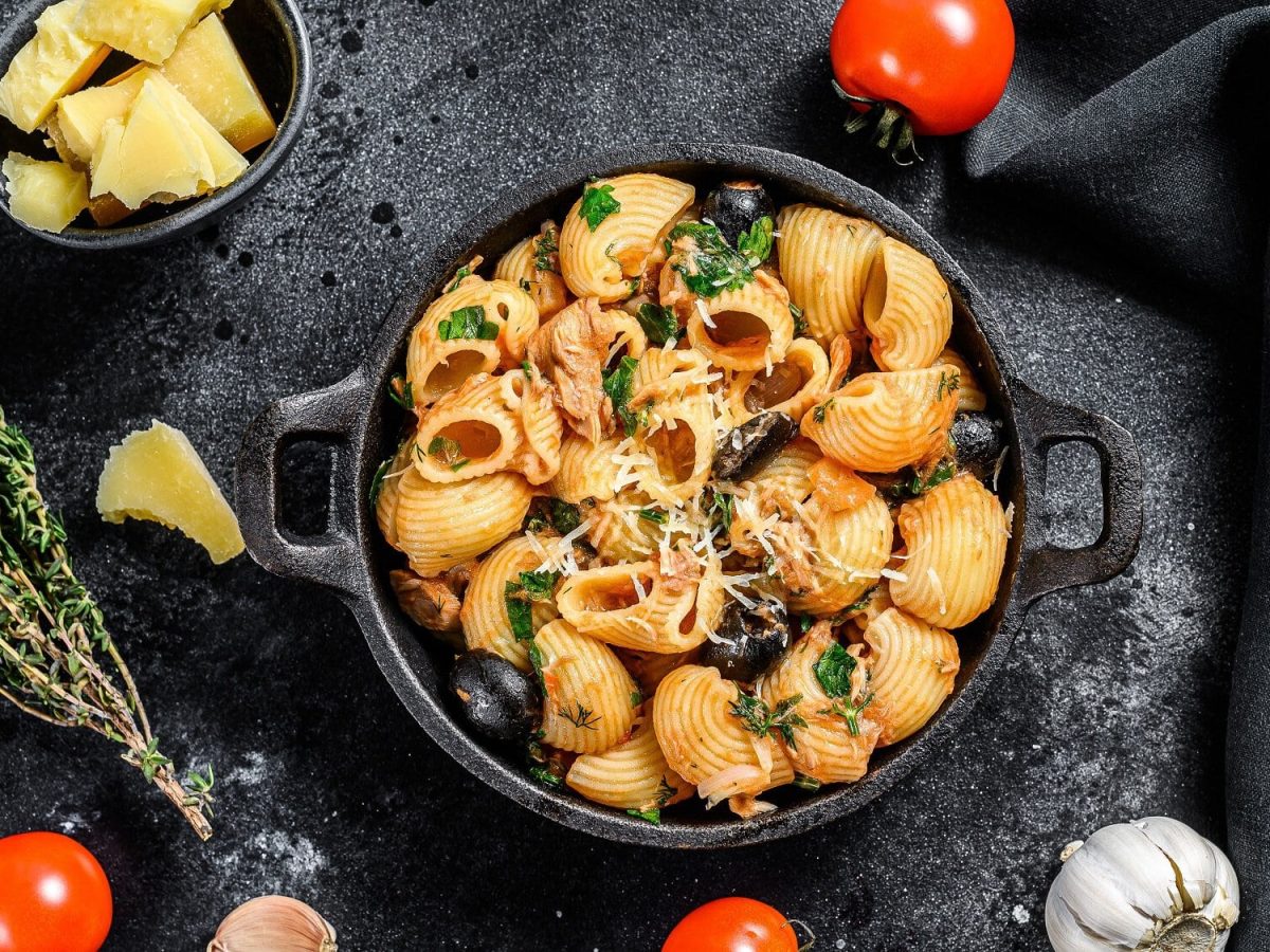 Draufsicht: dunkler Untergund, eine ofenfeste form mit Pasta in Tomaten-Kapern-Sauce. Drum herum liegen Gemüse und Kräuter.