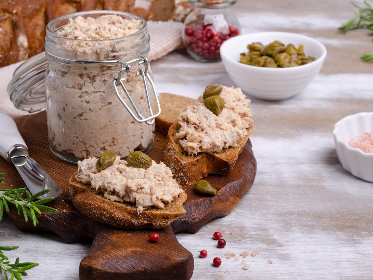 Frontal: Ein Einmachglas mit selbst gemachtem Lachs-Rillette steht auf einem dunklem Holzbrett. das liegt auf einem hellem Untergrund. Daneben lieben zwei kleine Weißbrotscheiben mit dem Lachs-Rille, Rosmarinzweige sowie eine kleine Schüssel mit Kapern.