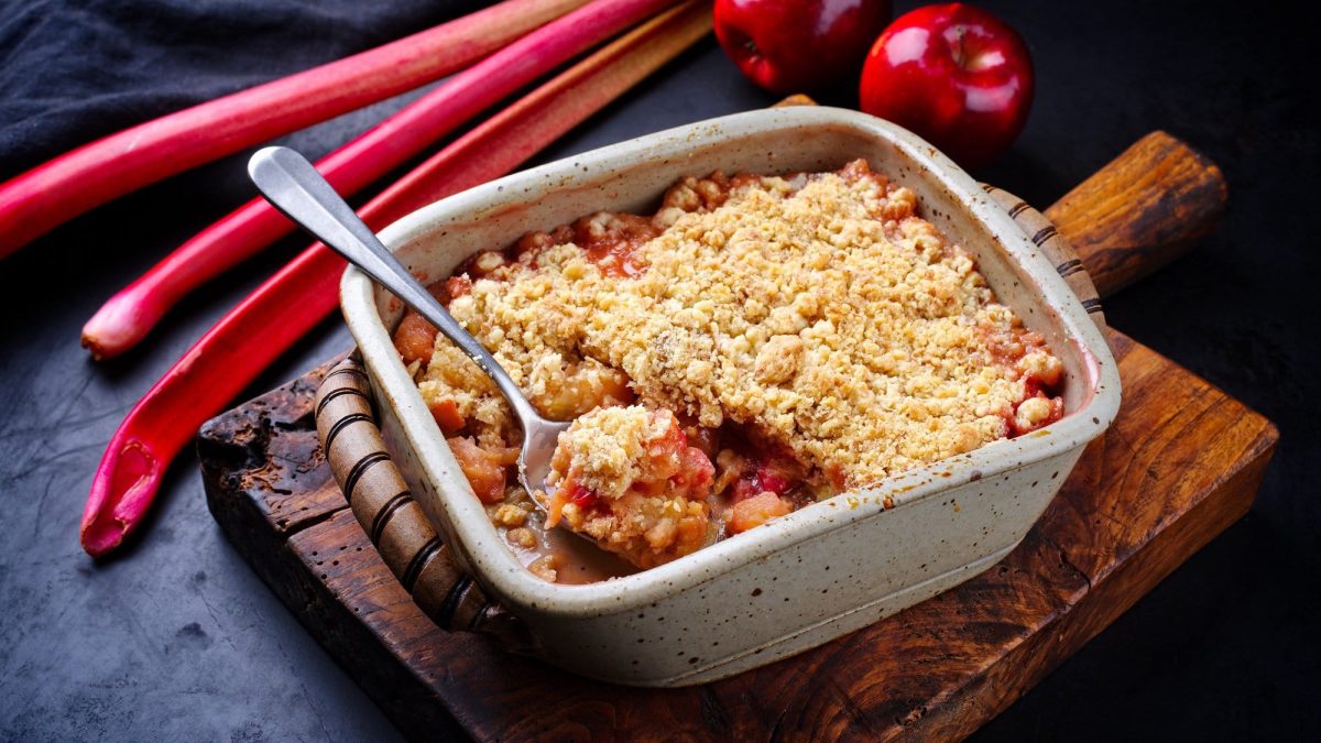 Eine Auflaufform mit Rhabarber-Crumble und einem Löffel darin auf einem Brett, daneben Rhabarber und Äpfel.