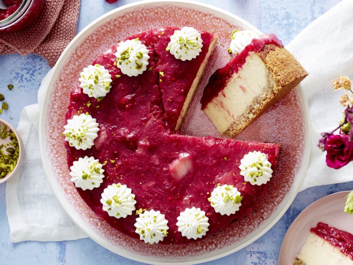 Draufsicht: Angeschnittener, runder Rhabarber-Grieß-Cheesecake mit Sahnetuffs. Daneben eine Schale mit zerbröselten Pistazien und Deko.