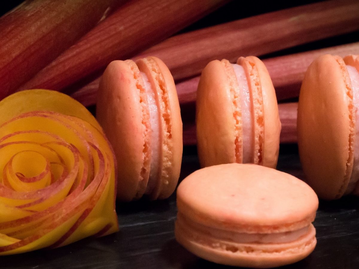 Vier gefüllte Macarons auf einem dunklen-schwarzen Hinter- und Untergrund mit Rhabarberstangen.