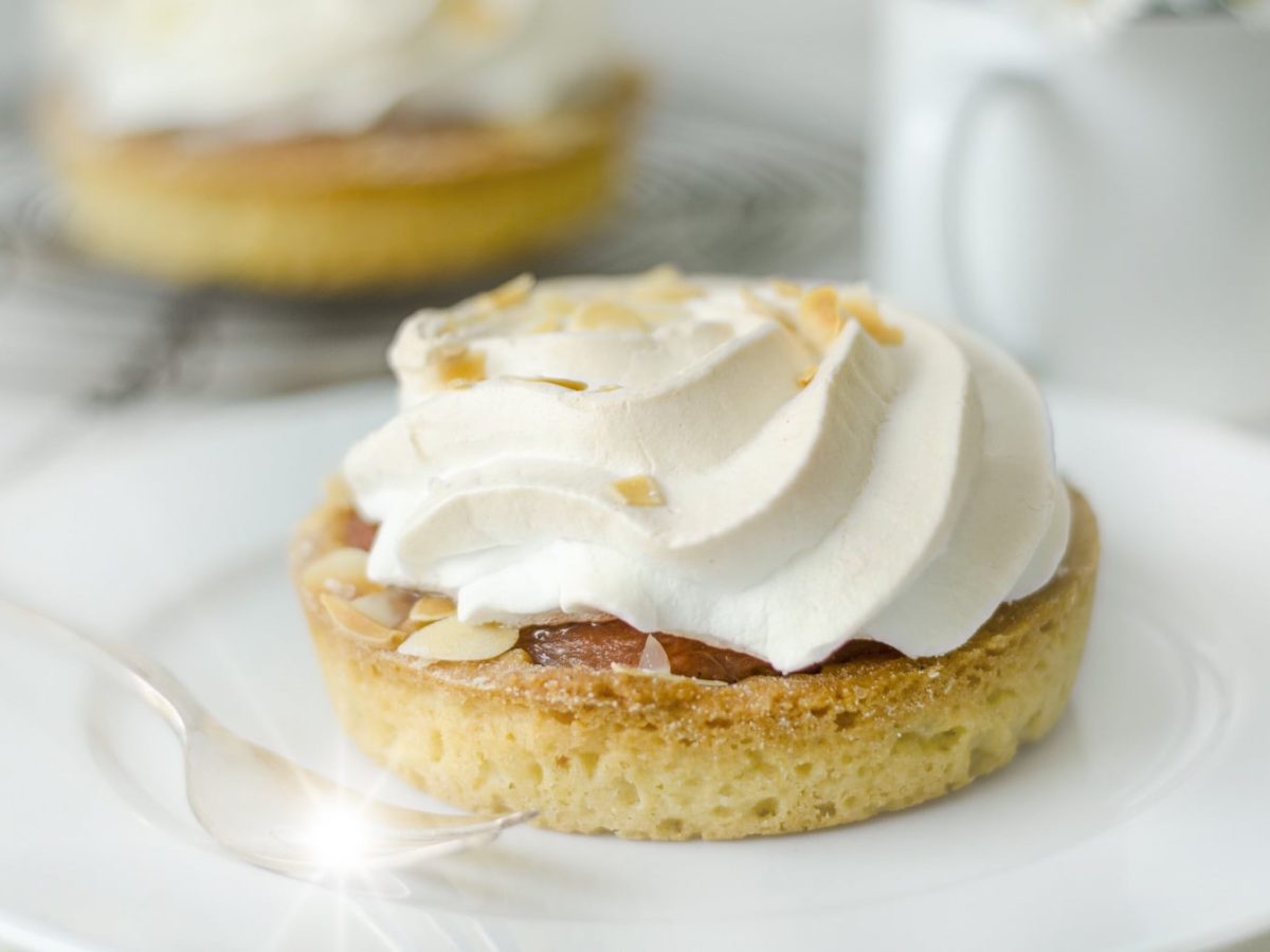 Ein weißer Teller mit der Rhabarber-Tartelette und der Meringue-Haube auf einem weißen Tisch.