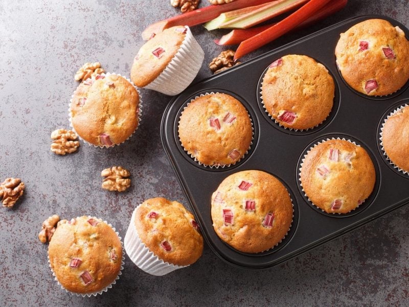 Ein Blech mit Rhabarber-Walnuss-Muffins in der Draufsicht, daneben liegen Walnusskerne, Rhabarberstangen und weitere Muffins.