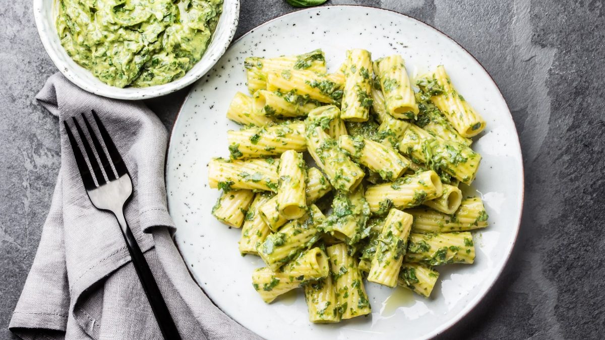 Ein weißer Teller mit den Rigatoni und Brokkoli-Joghurt-Sauce auf einem dunklen Steinuntergrund direkt von oben fotografiert. Daneben noch einige Basilikumblättchen.