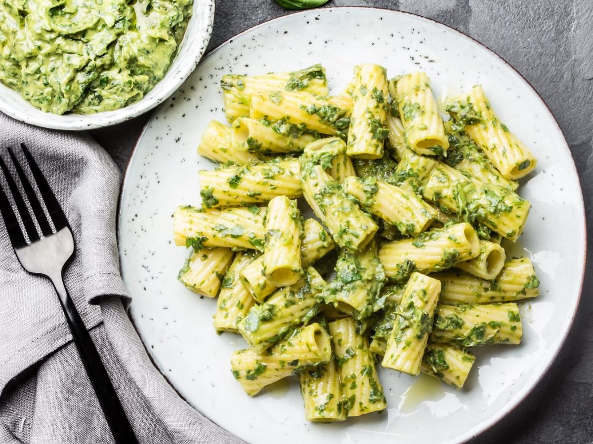 Ein weißer Teller mit den Rigatoni und Brokkoli-Joghurt-Sauce auf einem dunklen Steinuntergrund direkt von oben fotografiert. Daneben noch einige Basilikumblättchen.