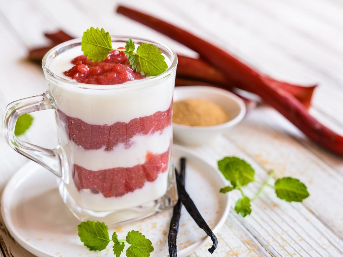 Ein Glas Schichtdessert mit Rhabarber und Kokoscreme, dekoriert mit Minze, dahinter Rhabarberstangen und weitere Minze.