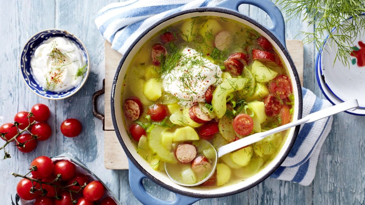 Draufsicht: Schmorgurken-Cabanossi-Eintopf mit saurer Sahne und Dill im Topf. Drum herum liegen Tomaten, Dill und saure Sahne vor hellblauem Hintergrund.