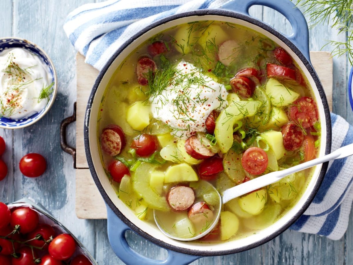 Draufsicht: Schmorgurken-Cabanossi-Eintopf mit saurer Sahne und Dill im Topf. Drum herum liegen Tomaten, Dill und saure Sahne vor hellblauem Hintergrund.