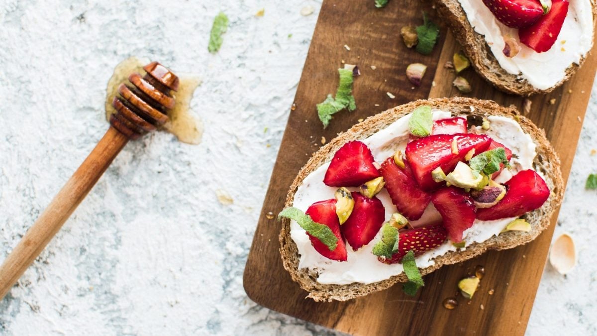 Draufsicht: Auf einem hellen Untergrund liegt ein Holzbrettchen mit zwei Vollkornbroten mit Erdbeer-Ricotta. Daneben liegt ein Holzlöffel mit Honig.