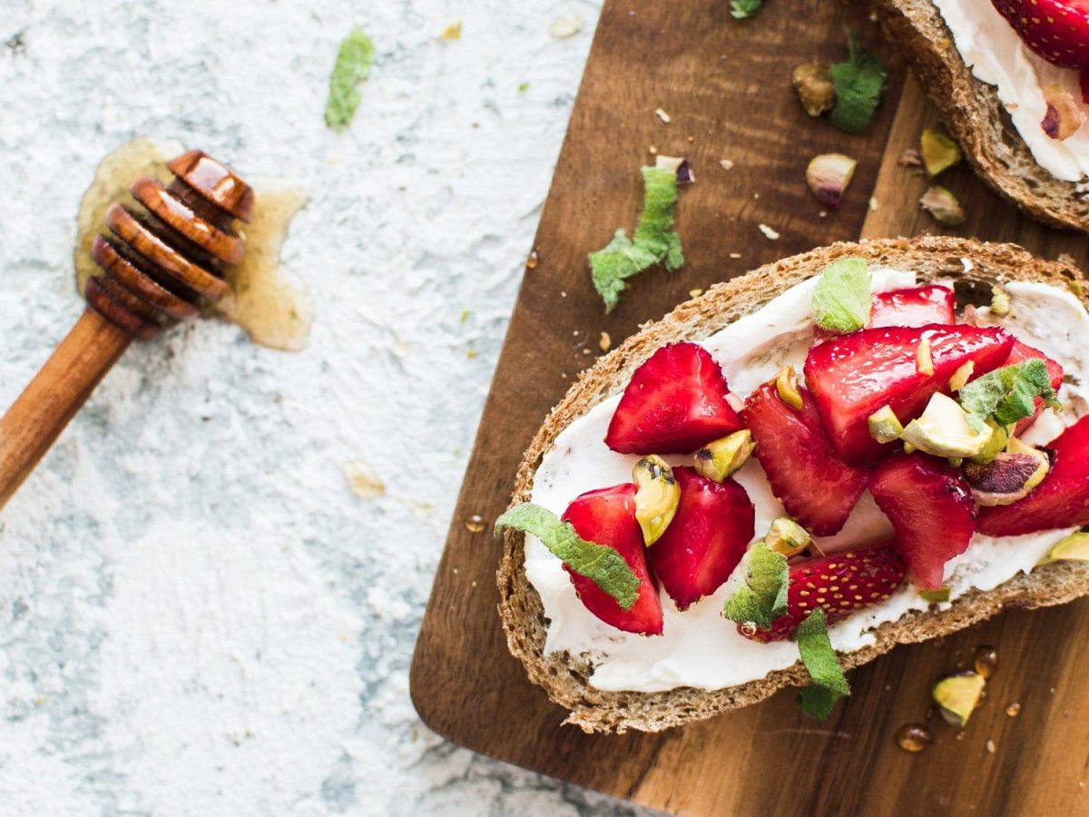 Draufsicht: Auf einem hellen Untergrund liegt ein Holzbrettchen mit zwei Vollkornbroten mit Erdbeer-Ricotta. Daneben liegt ein Holzlöffel mit Honig.