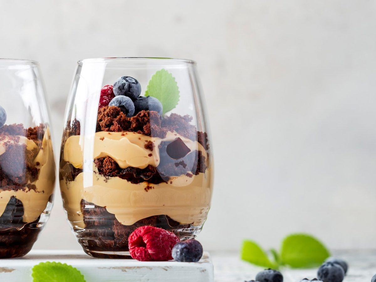 Zwei Gläser mit dem Schoko-Beeren-Dessert auf einem hellen Tisch mit Heidelbeeren und Zitronenmelisse.