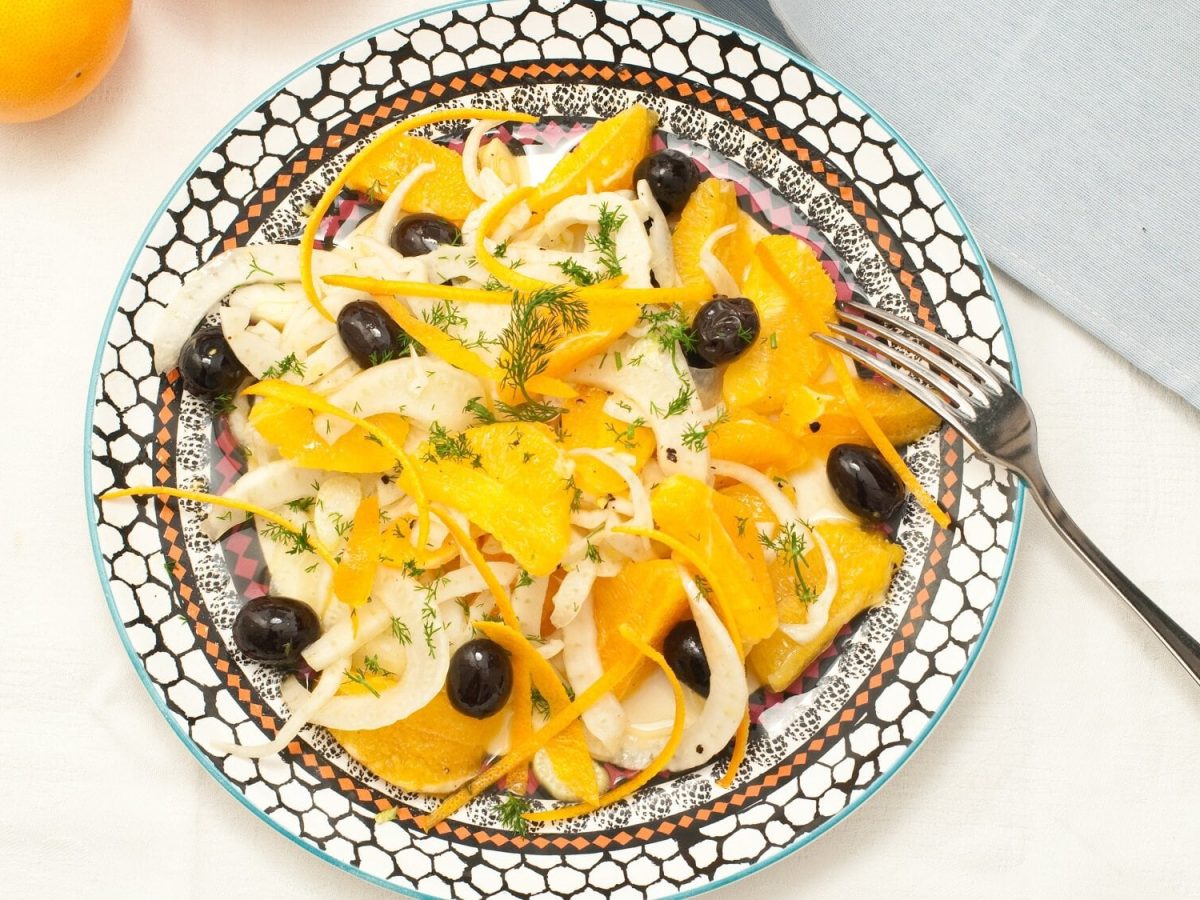 Draufsicht: ein mediterran bemalter Teller mit einem sizilianischem Fenchel-Oliven-Salat mit Mandel-Dressing steht auf einem weißen Untergrund. Daneben liegen eine Gabel und ein hellblaues Tuch.