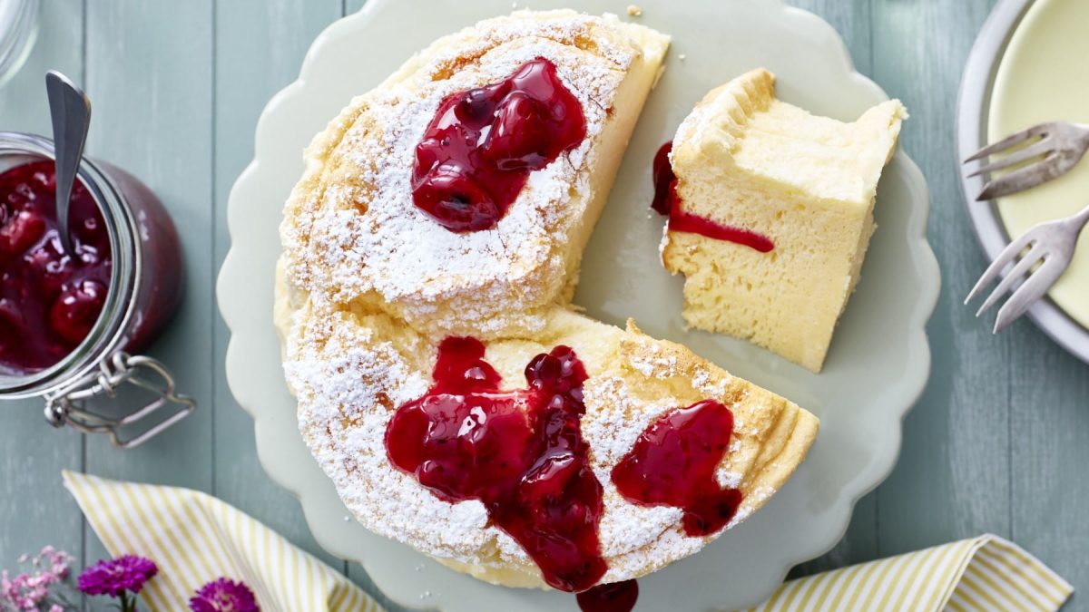 Angeschnittener Soufflé-Käsekuchen mit Rote Grütze auf einem weißen Teller, drum herum stehen ein Glas Grütze, Servietten, Gabeln, Geschirr und lila Blumen.