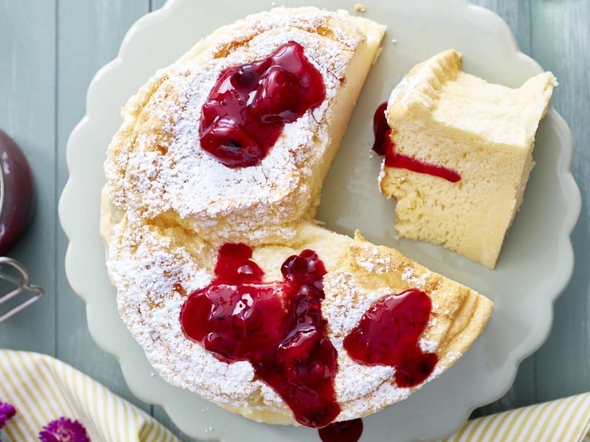 Angeschnittener Soufflé-Käsekuchen mit Rote Grütze auf einem weißen Teller, drum herum stehen ein Glas Grütze, Servietten, Gabeln, Geschirr und lila Blumen.