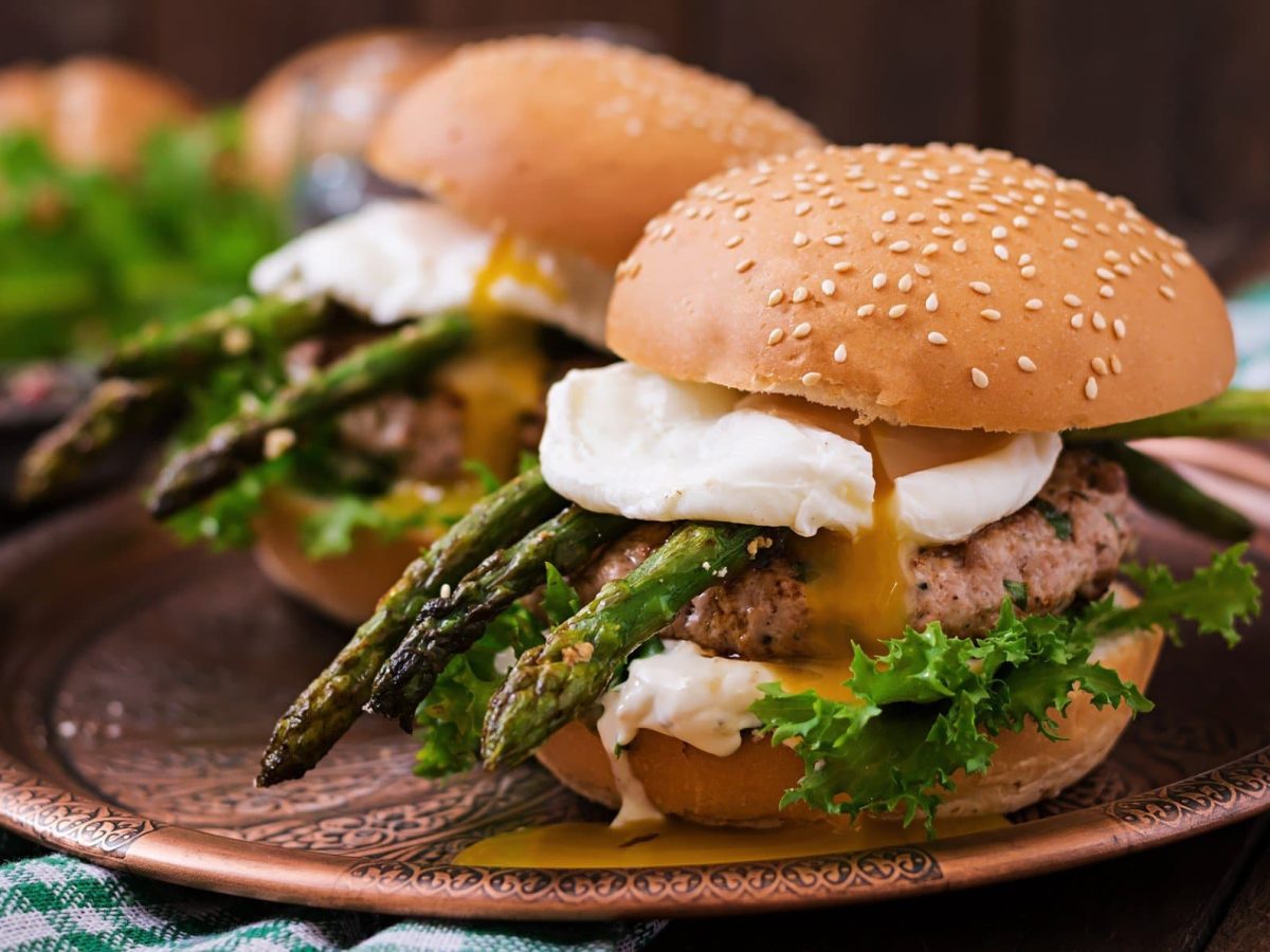 Zwei Spargel-Burger mit pochiertem Ei, Chicken-Patty, grünem Spargel und Burrata auf braunem Teller. Im Hintergrund weitere Zutaten. Frontalansicht.