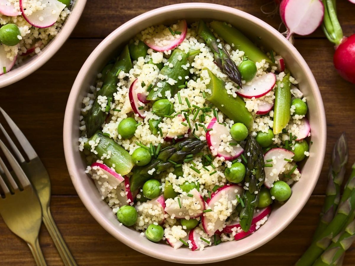 Zwei Schüsseln Spargel-Couscous-Salat mit Erbsen und Radieschen in der Draufsicht, daneben zwei Gabeln, grüne Spargelstangen und Radieschen.