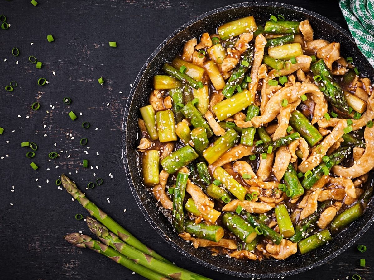 Eine schwarze Pfanne mit Spargel und Hähnchenstreifen auf einem schwarzen Untergrund. Daneben Sesam, Frühlingslauchringe und drei Stangen grüner Spargel. Direkt von oben fotografiert.