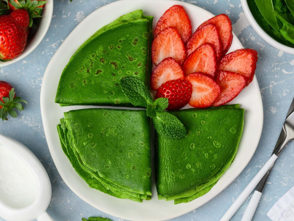 Draufsicht: auf einem blauen Untergrund steht ein Teller mit Spinat-Minze-Pfannkuchen mit Erdbeeren. Drumherum stehen eine Schale mit Erdbeeren, aufgeschnittenen Erdbeeren, ein Kännchen mit einer weißen Flüssigkeit sowie Messer und Gabel.