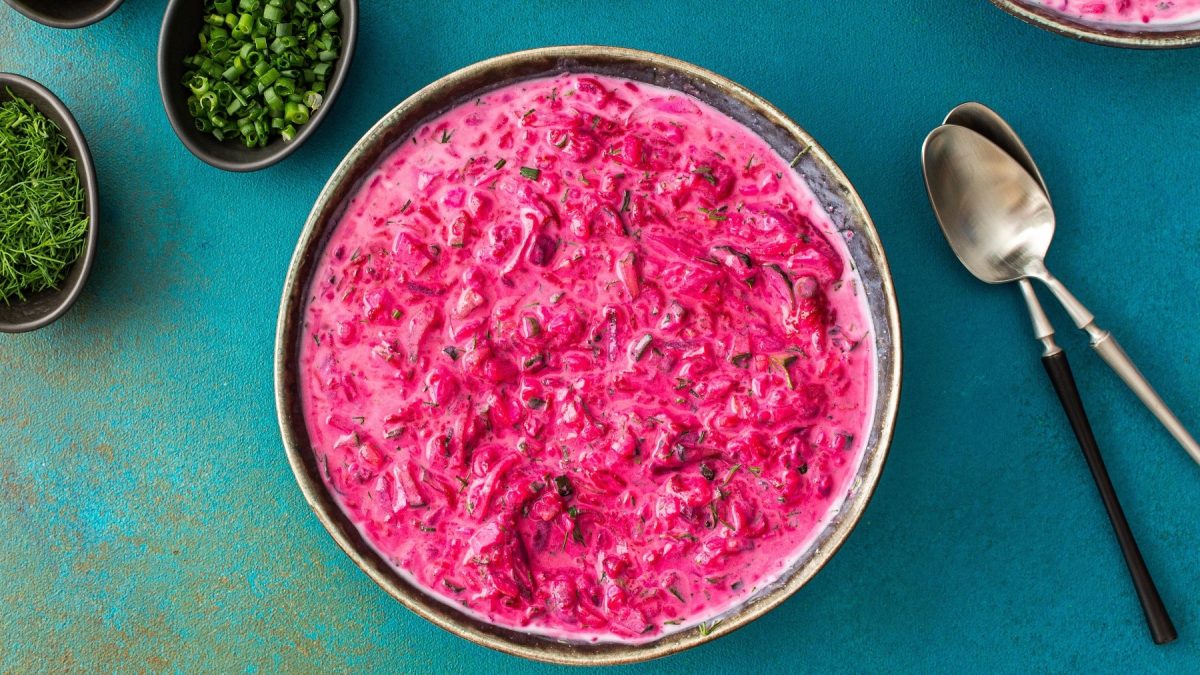 Eine Schale mit Svekolnik (pinke Suppe) auf einem kontrastreichen blauen Tisch mit Kräutern und zwei Löffeln.