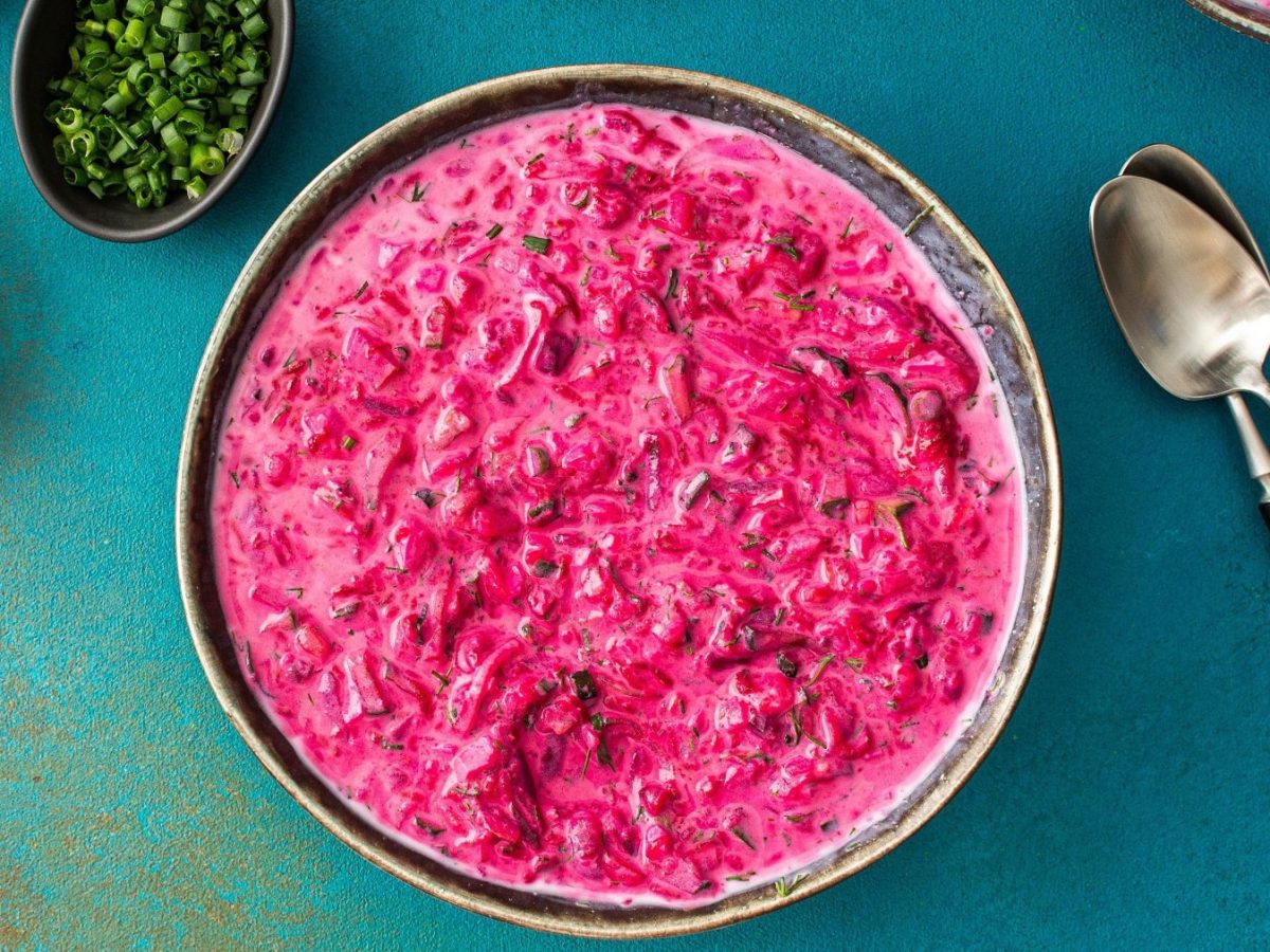 Eine Schale mit Svekolnik (pinke Suppe) auf einem kontrastreichen blauen Tisch mit Kräutern und zwei Löffeln.