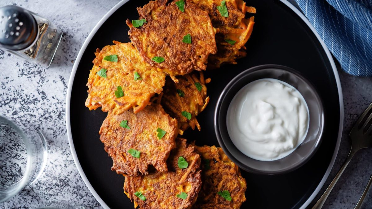 Sweet Potato Fritters mit weißem Dip auf schwarzem Teller und grauem Untergrund. Im Bild Salzstreuer. Draufsicht.