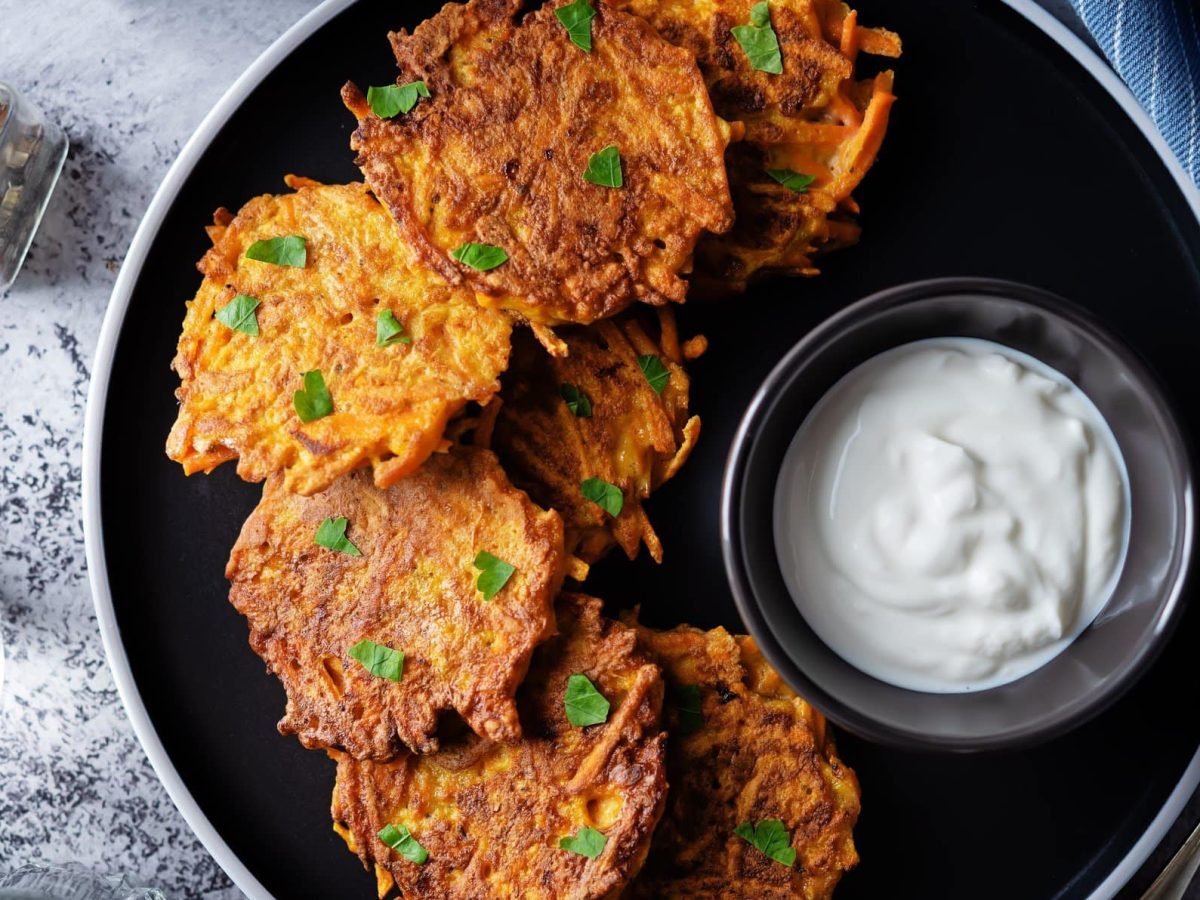 Sweet Potato Fritters mit weißem Dip auf schwarzem Teller und grauem Untergrund. Im Bild Salzstreuer. Draufsicht.