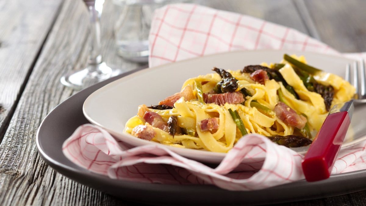 Tagliatelle Carbonara mit violettem Spargel auf Pasta-Teller, Tuch und Unterteller auf Holztisch. Daneben Weinglas mit Rotwein. Draufsicht.