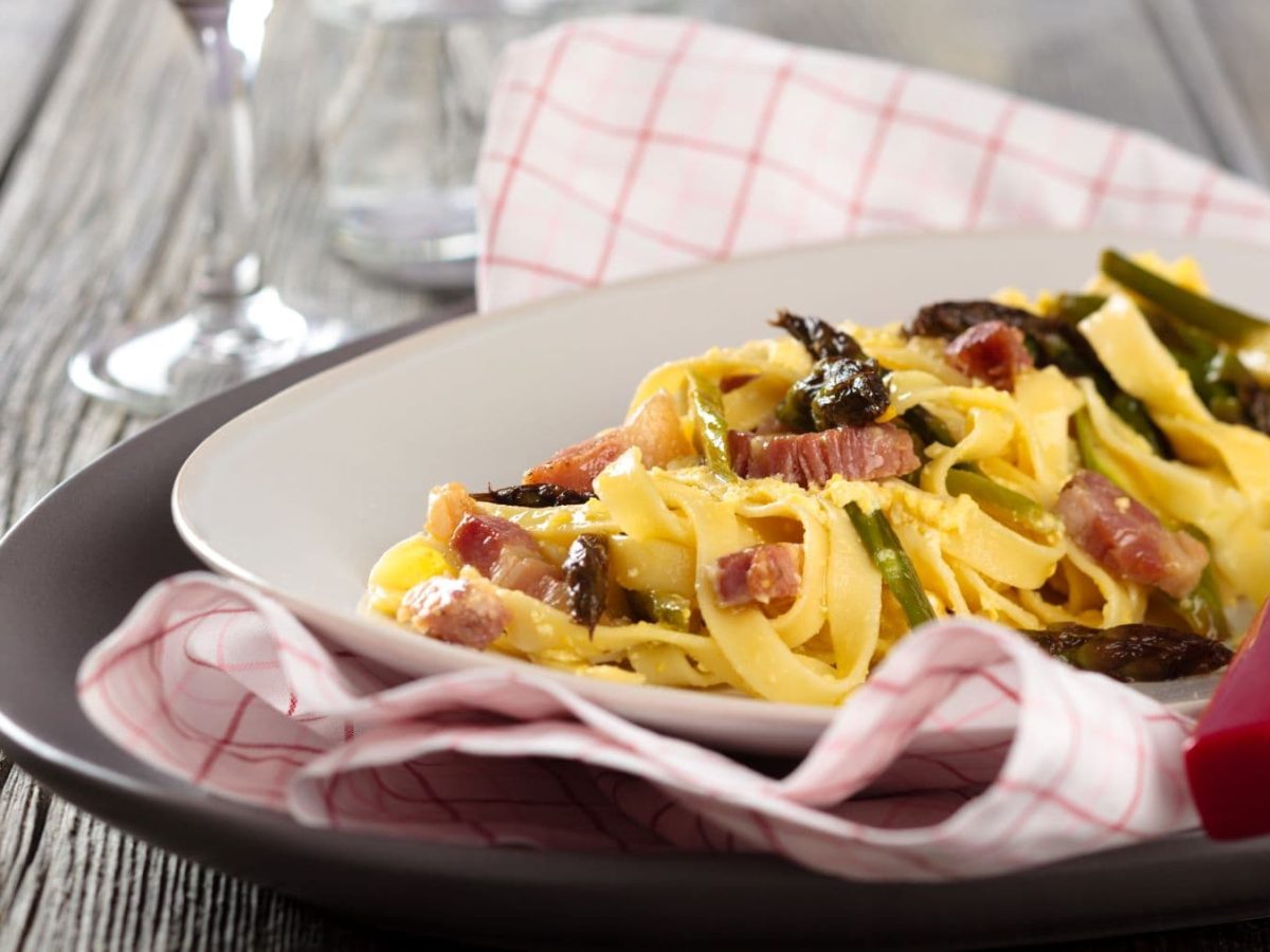 Tagliatelle Carbonara mit violettem Spargel auf Pasta-Teller, Tuch und Unterteller auf Holztisch. Daneben Weinglas mit Rotwein. Draufsicht.