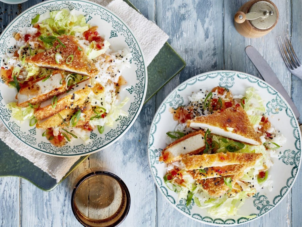 Zwei Teller mit angeschnittenen Tofu-Schnitzel mit Chinakohl neben Gewürzmühle, Glas und Besteck auf hellblauem Holzuntergrund.