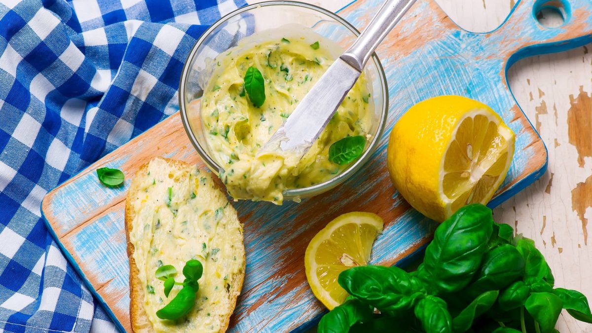 Eine Schale Wodka-Butter mit Basilikum, daneben eine Scheibe Brot, die mit der Butter bestrichen ist, außerdem eine Zitrone und frischer Basilikum. Alles Draufsicht.