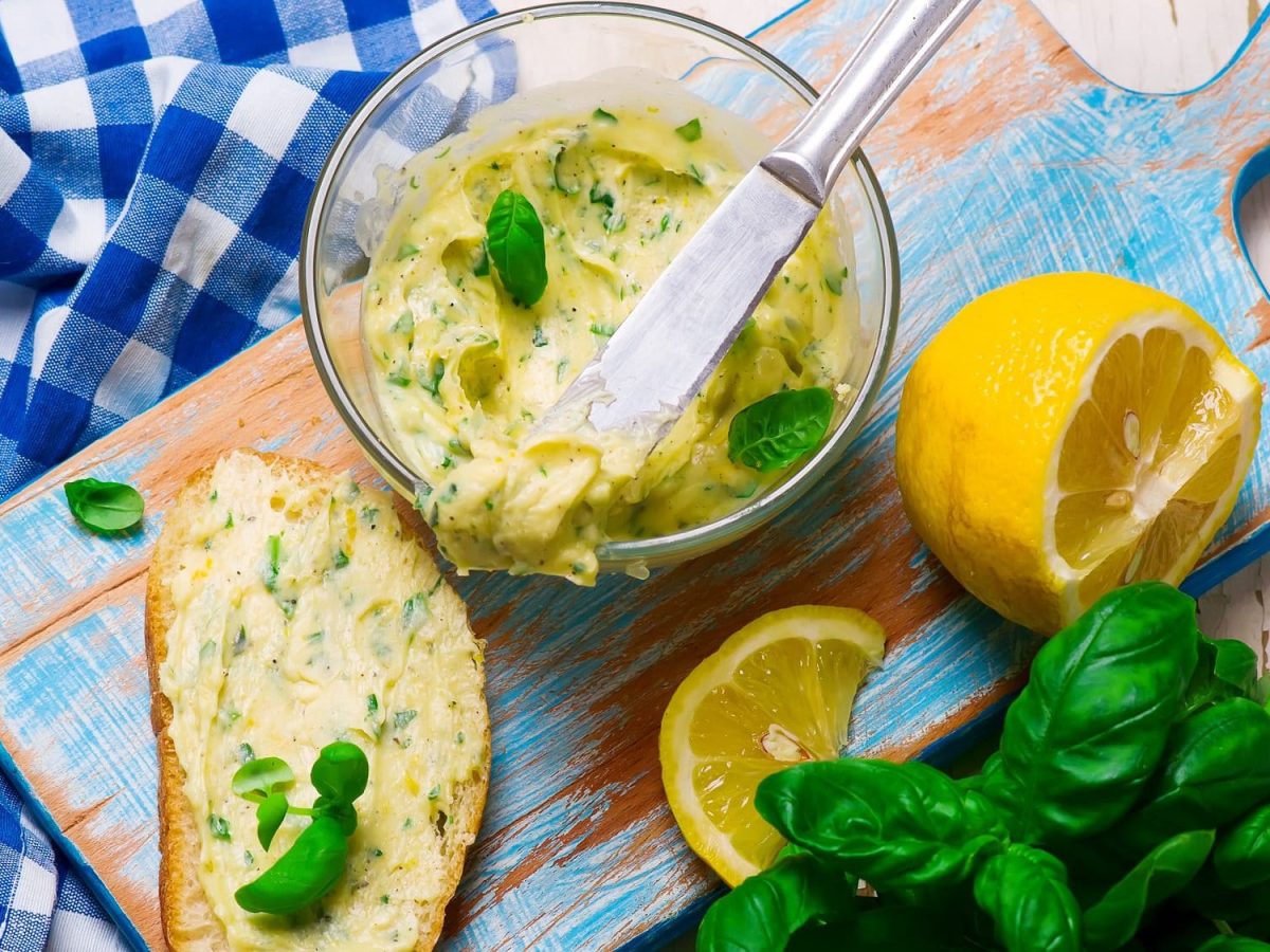 Eine Schale Wodka-Butter mit Basilikum, daneben eine Scheibe Brot, die mit der Butter bestrichen ist, außerdem eine Zitrone und frischer Basilikum. Alles Draufsicht.