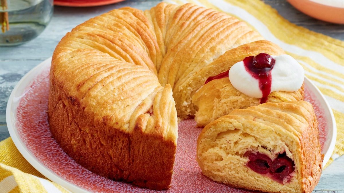 Angeschnittener Wollknäuel-Butterkuchen mit Kirschen und Sahne auf einem rosa Teller.