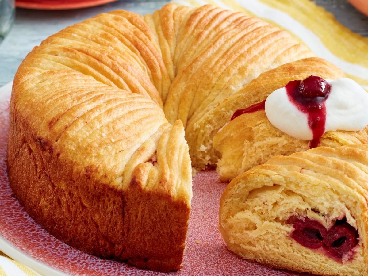 Angeschnittener Wollknäuel-Butterkuchen mit Kirschen und Sahne auf einem rosa Teller.