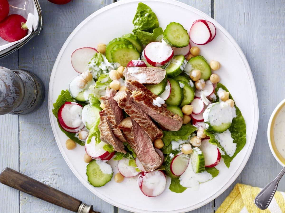 Draufsicht: Zaziki-Salat mit Steakstreifen auf einem weißen Teller. Außerdem eine Schüssel mit weißer Sauce, Gewürzmühle und Radieschen vor hellem Holzhintergrund.