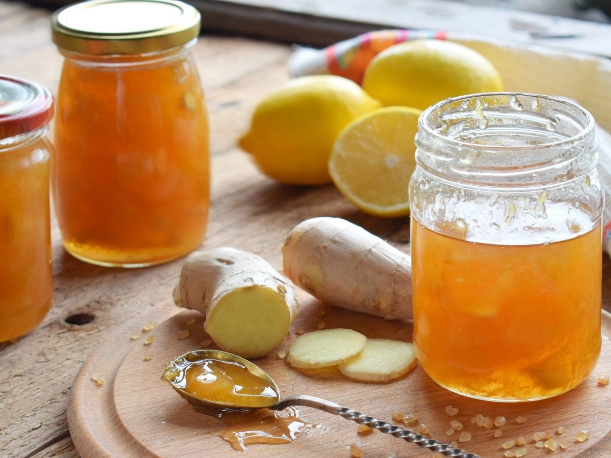 Frontal: drei Gläser Zitronen-Ingwer-Marmelade stehen auf einem Untergrund aus Holz. Drum herum liegen frischer Ingwer und Zitronen und ein Teelöffel.